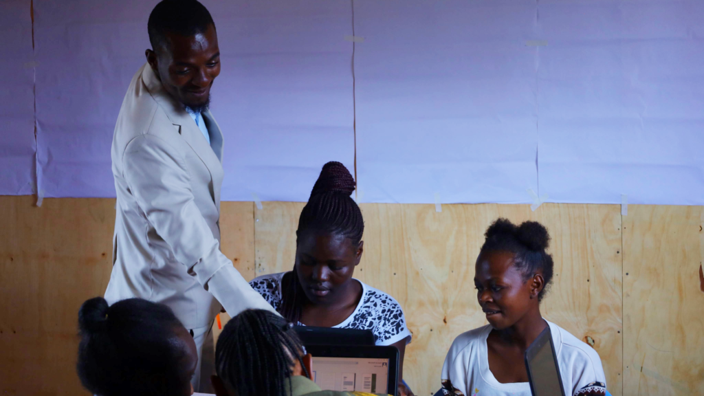 Founder Sain Ramadhan teaches the first class at Kua Digital's new partner project, A Pack A Month. Sain Ramadhan is standing next to two sitting students and is pointing at their laptop.