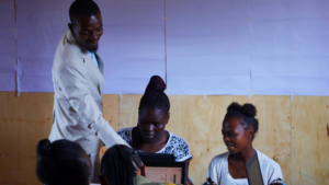 Founder Sain Ramadhan teaches the first class at Kua Digital's new partner project, A Pack A Month. Sain Ramadhan is standing next to two sitting students and is pointing at their laptop.